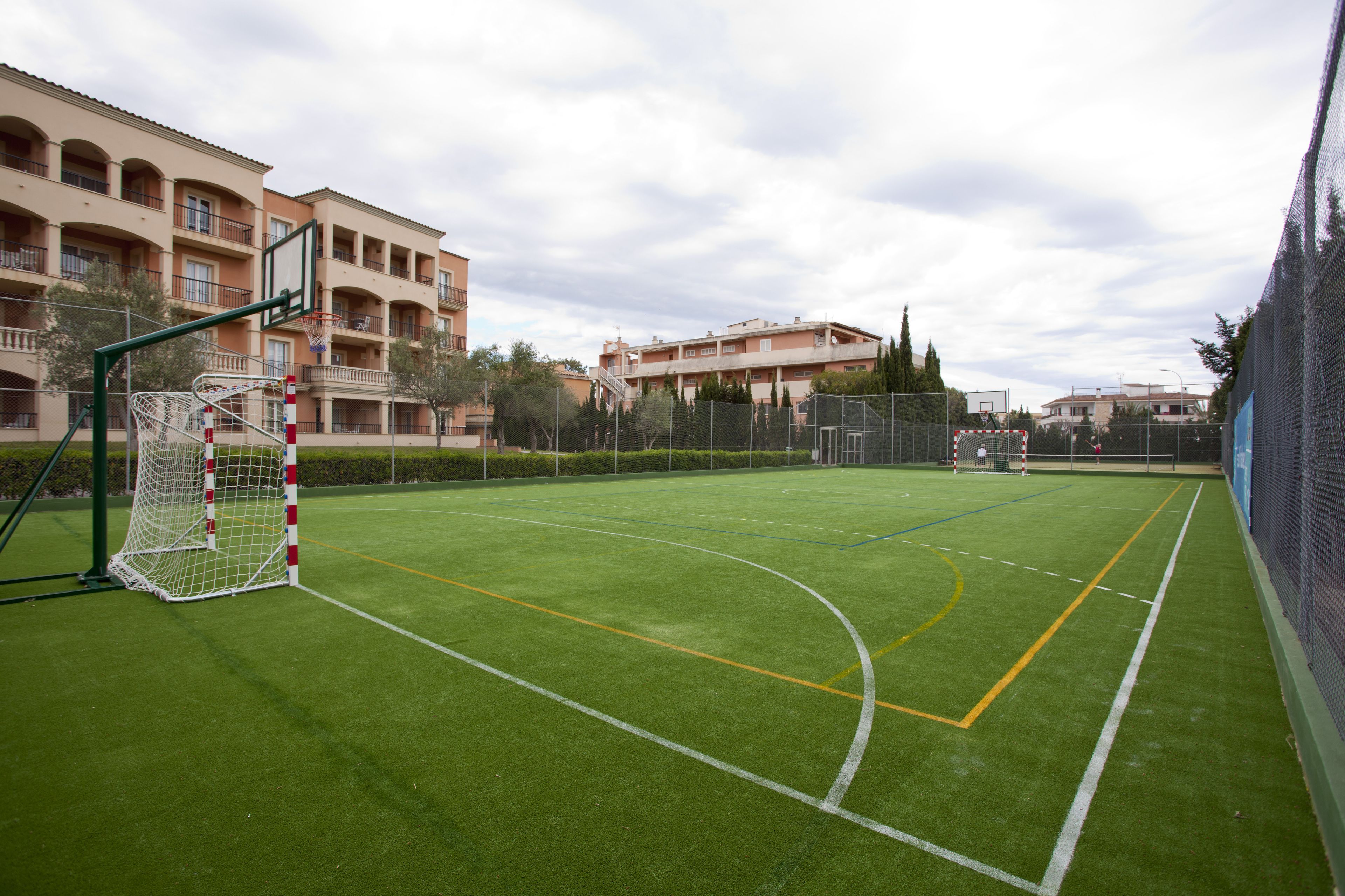 Una cancha deportiva con un poste de portería, rodeada por una valla y edificios al fondo.
