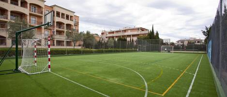 Una cancha deportiva con un poste de portería, rodeada por una valla y edificios al fondo.