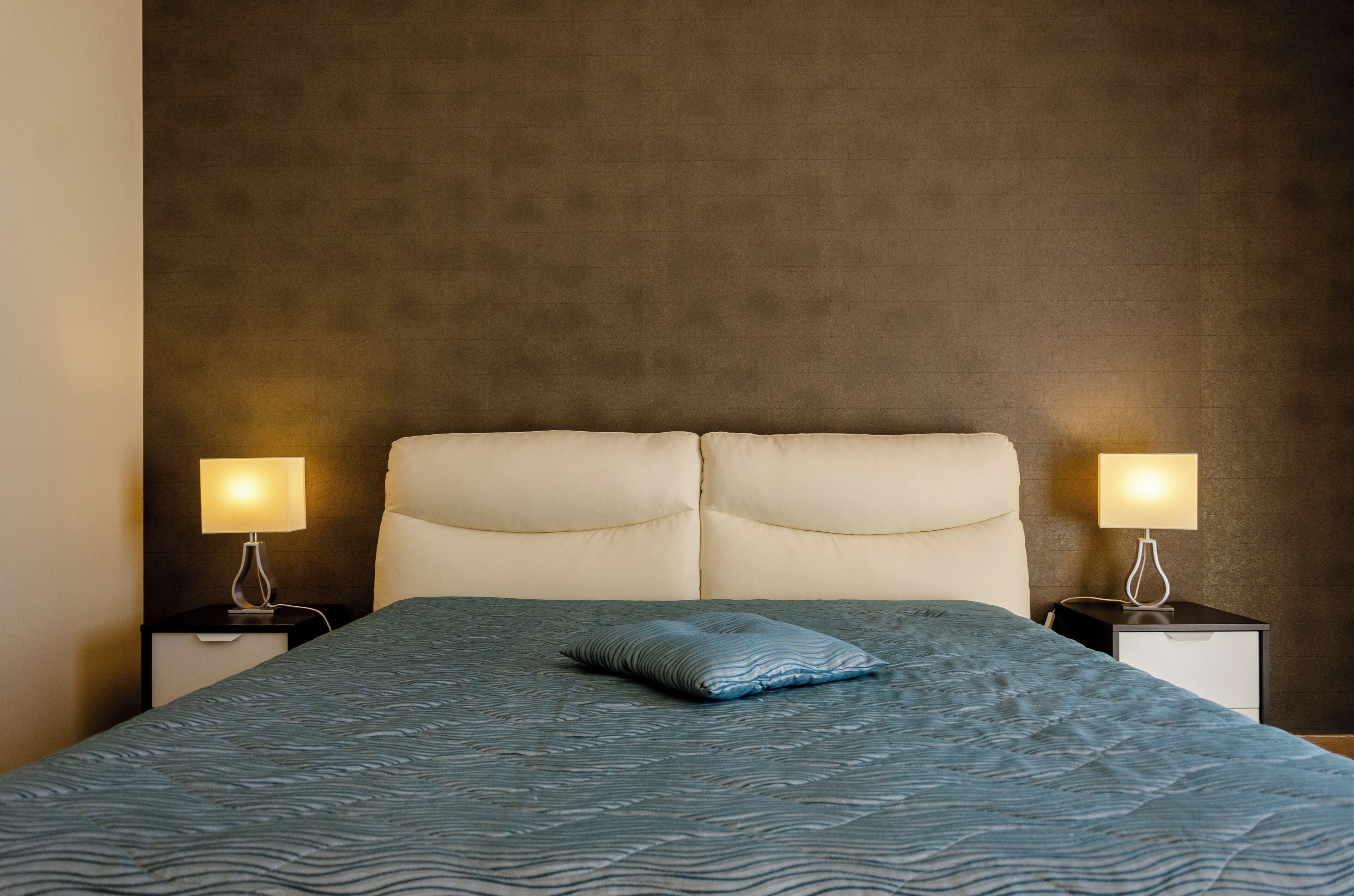 Een netjes opgemaakt bed met een blauw geborreld sprei en één kussen, met aan beide kanten nachtkastjes met lampen.