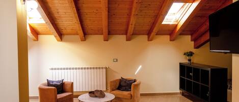 Una habitación con vigas de madera en el techo, un radiador, un mueble negro para televisión y dos sillones con una mesita.
