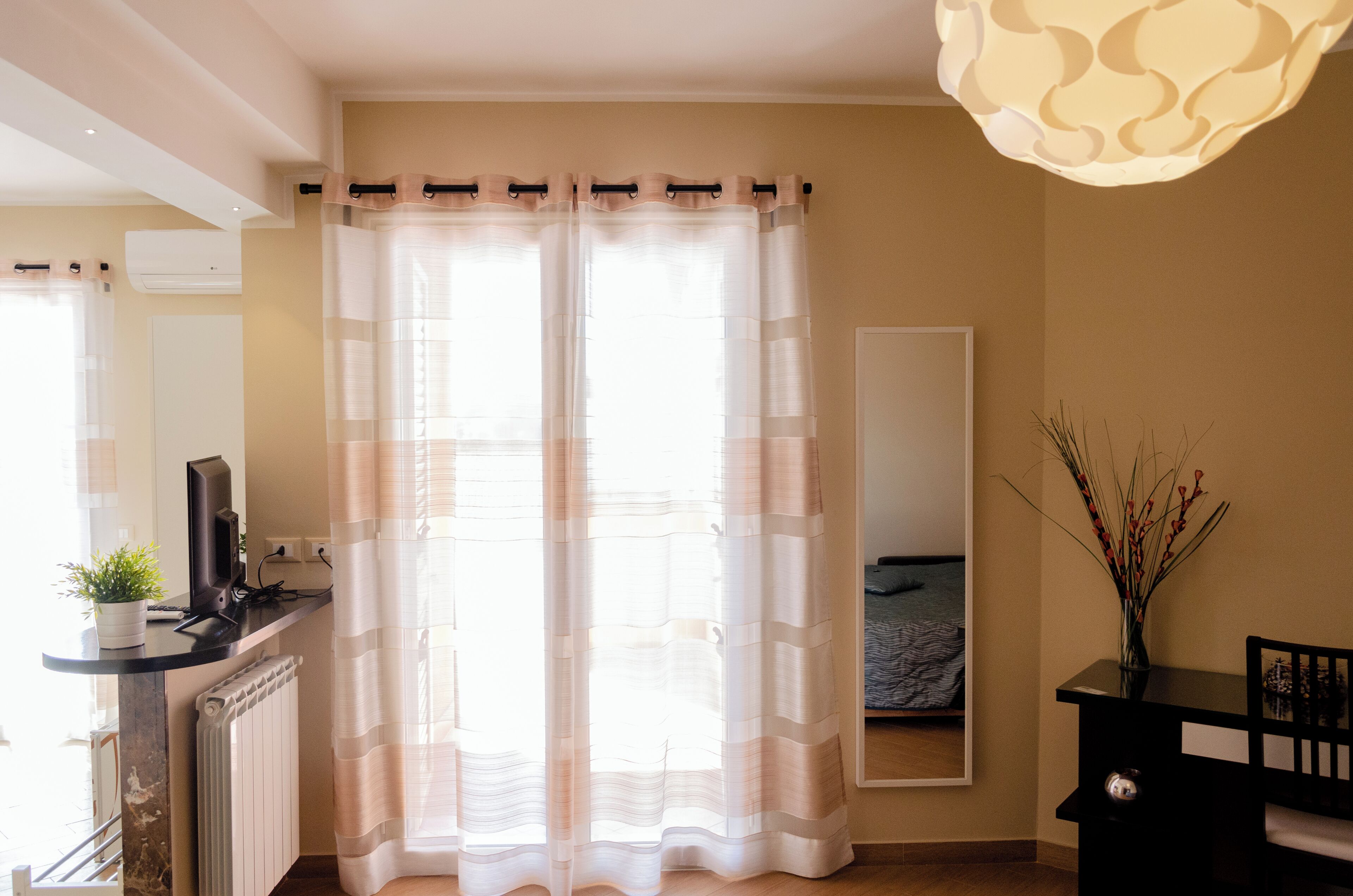 Una habitación con cama, escritorio con computadora, silla, espejo y ventana con cortinas traslúcidas.