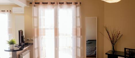 Una habitación con cama, escritorio con computadora, silla, espejo y ventana con cortinas traslúcidas.