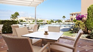 Un patio con mobiliario de mimbre, una mesa y vistas a una piscina y edificios al fondo.