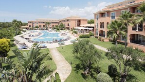 Un resort con piscina central rodeada de sillas de descanso y sombrillas, varias edificaciones con balcones y césped y árboles bien cuidados.
