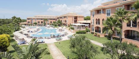 Un complexe hôtelier doté d’une piscine centrale entourée de chaises longues et de parasols, de plusieurs bâtiments avec des balcons, ainsi que de pelouses et d’arbres bien entretenus.