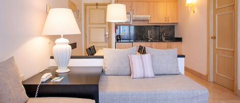 A hotel room with a bed, a sofa, a desk with a lamp, a phone, and a kitchenette in the background.