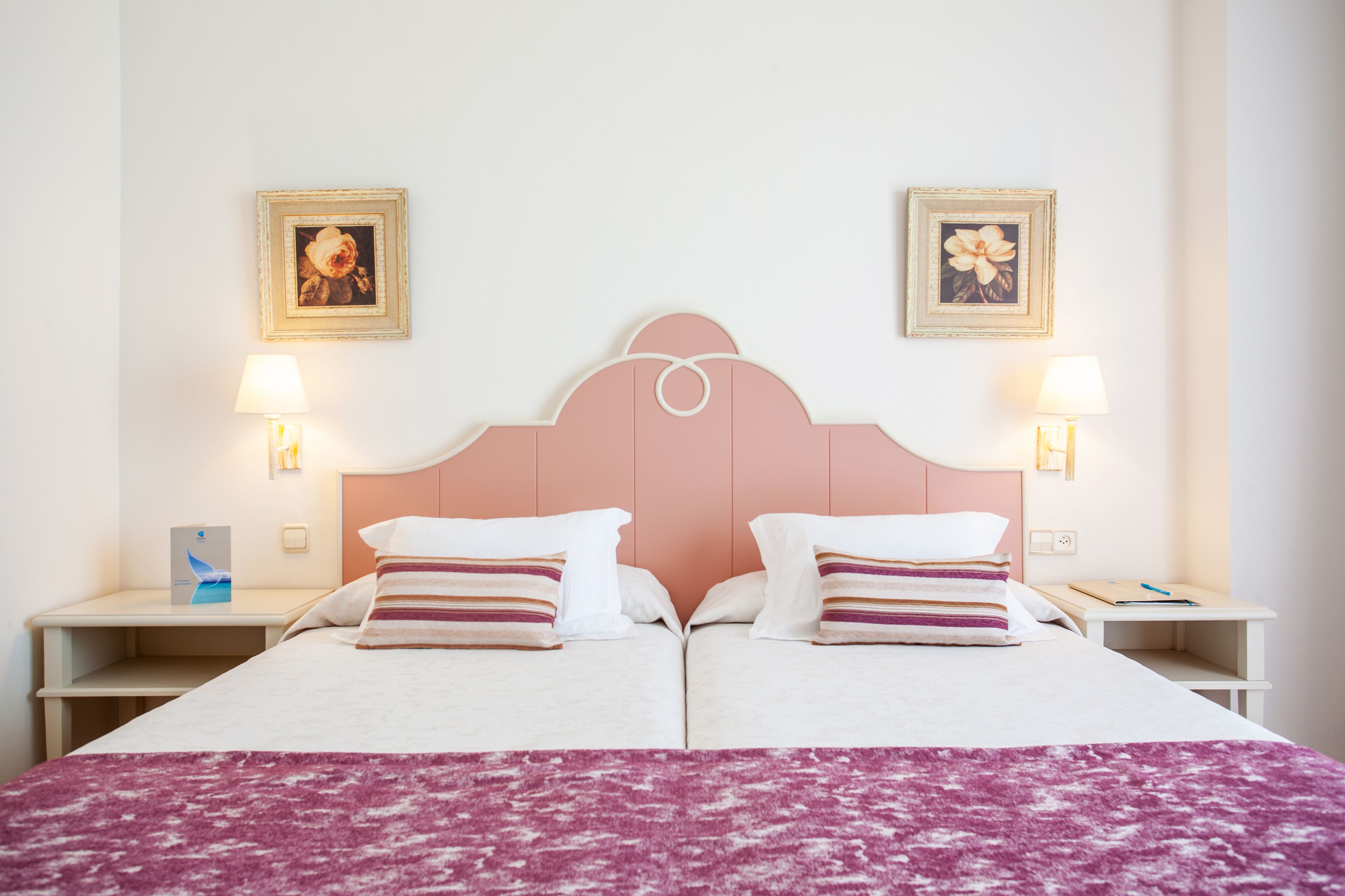 Un lit double avec une tête de lit rose, deux tables de chevet avec des lampes et deux tableaux encadrés accrochés au mur.