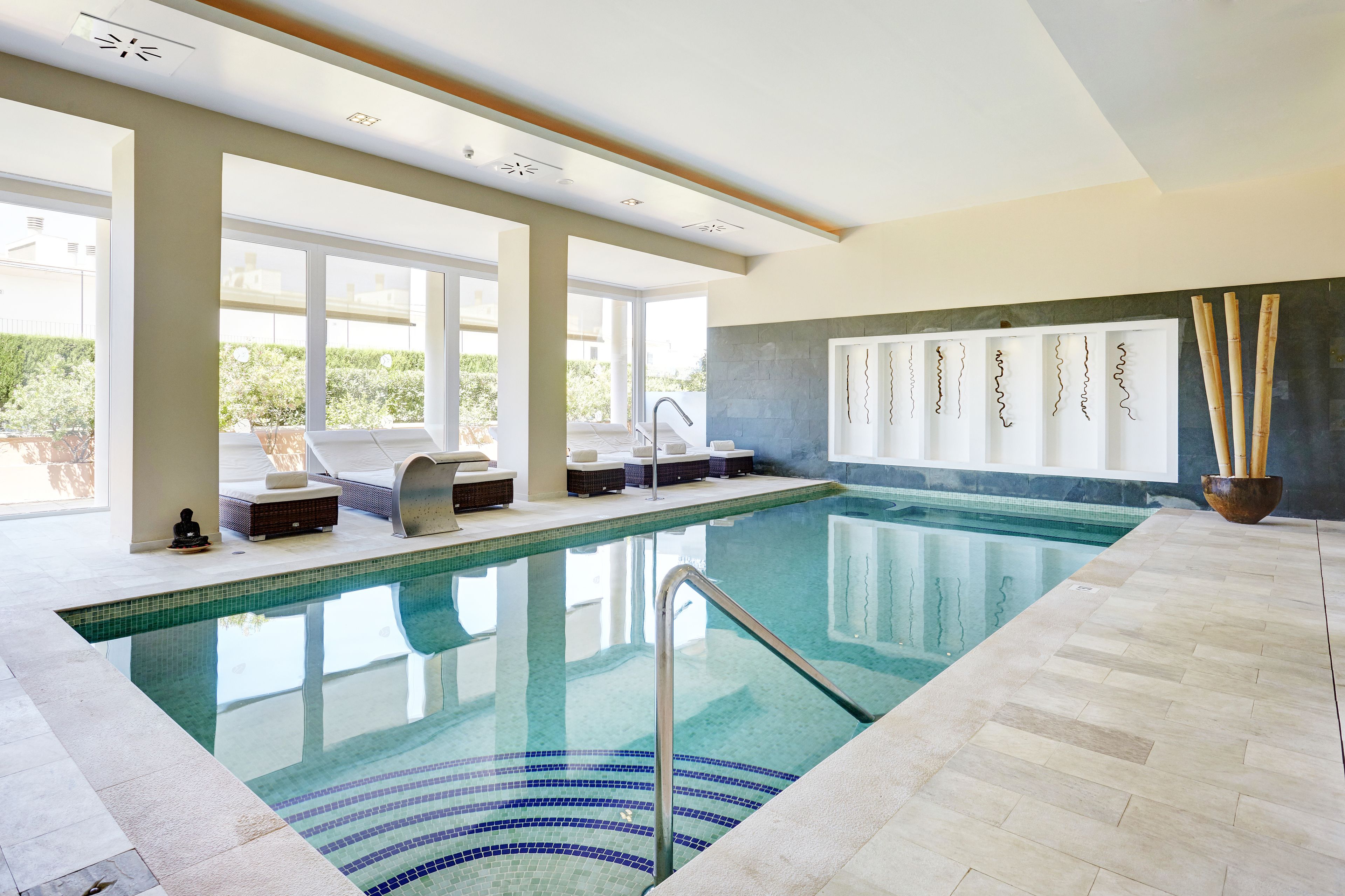 Una piscina interior con paredes de vidrio transparente, suelo de baldosas y una zona de estar con cojines y una pequeña estatua de Buda.