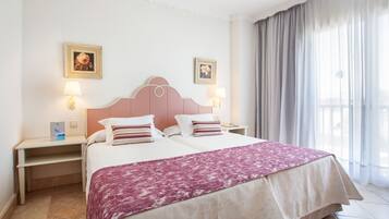 A double bed with a pink headboard, two bedside tables with lamps, a decorative pillow, and a window with curtains.