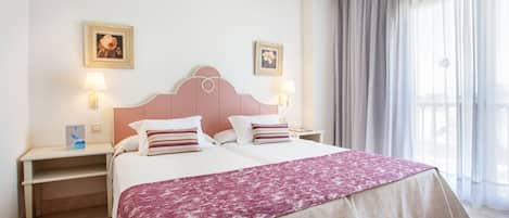 A double bed with a pink headboard, two bedside tables with lamps, a decorative pillow, and a window with curtains.