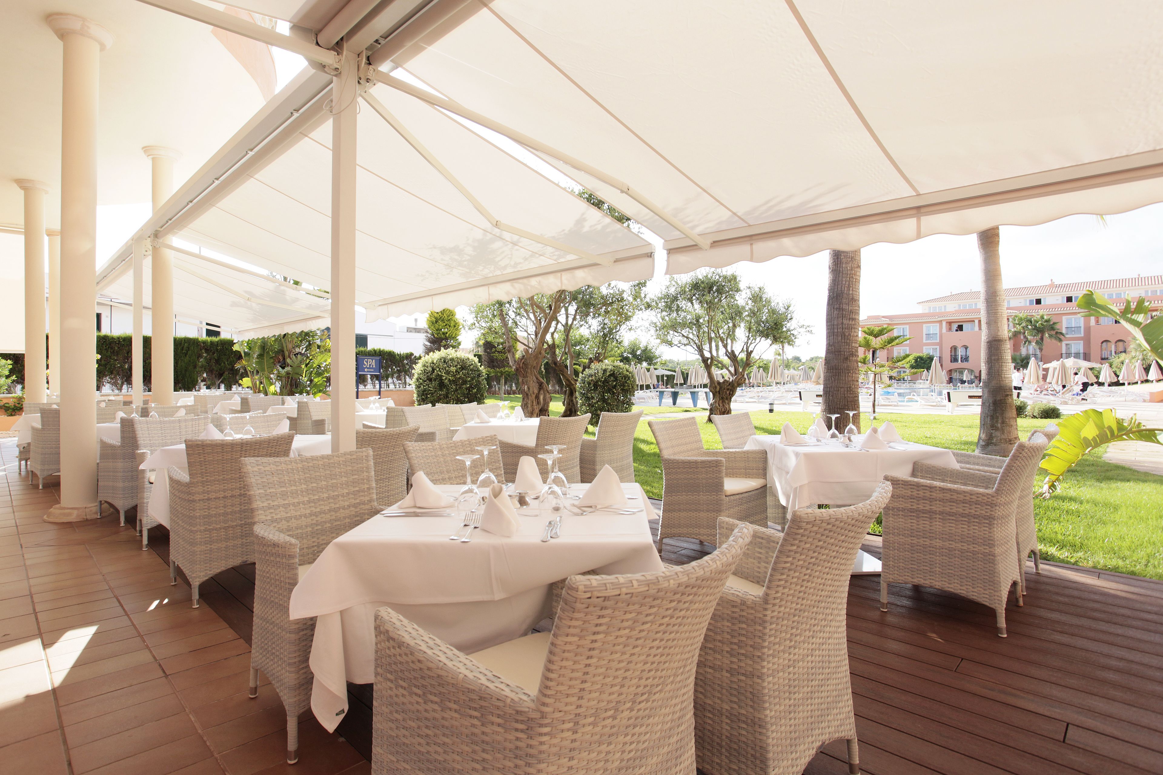 An outdoor dining area with wicker chairs and tables, covered by a white canopy, set against a backdrop of greenery and a building.