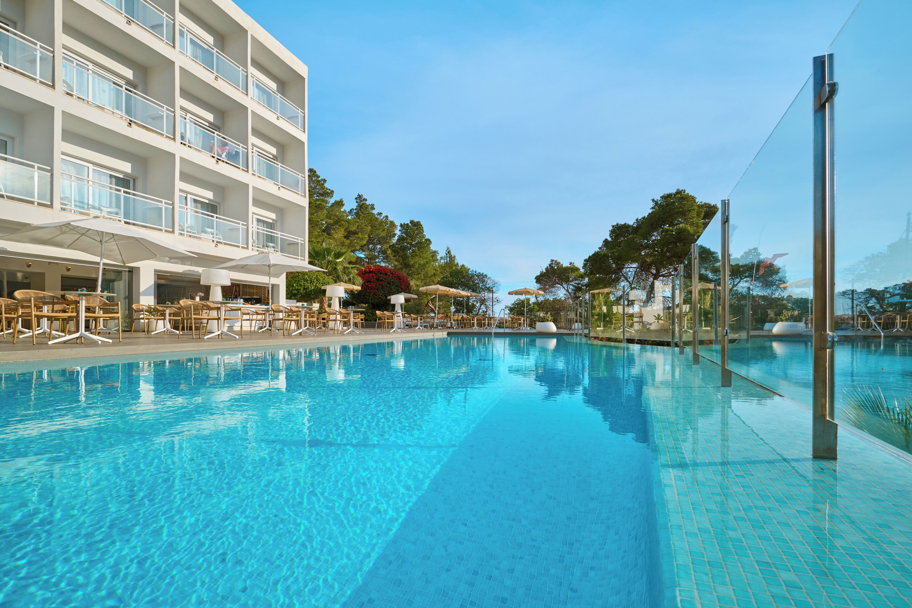 A swimming pool with lounge chairs and umbrellas, a multi-story building with balconies, and trees in the background.