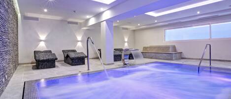 A modern indoor swimming pool with a blue water feature, seating areas, and a skylight.