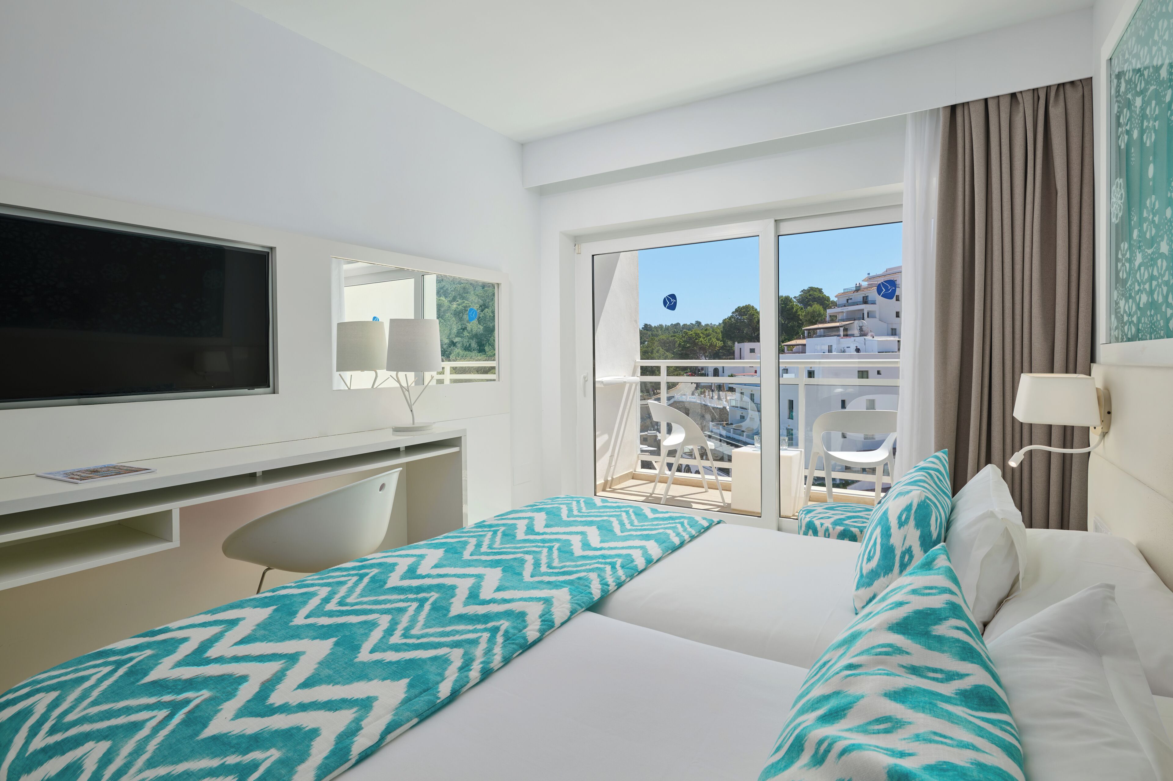 Habitació d'hotel amb llit, escriptori, cadira, televisió i balcó amb vista a edificis.