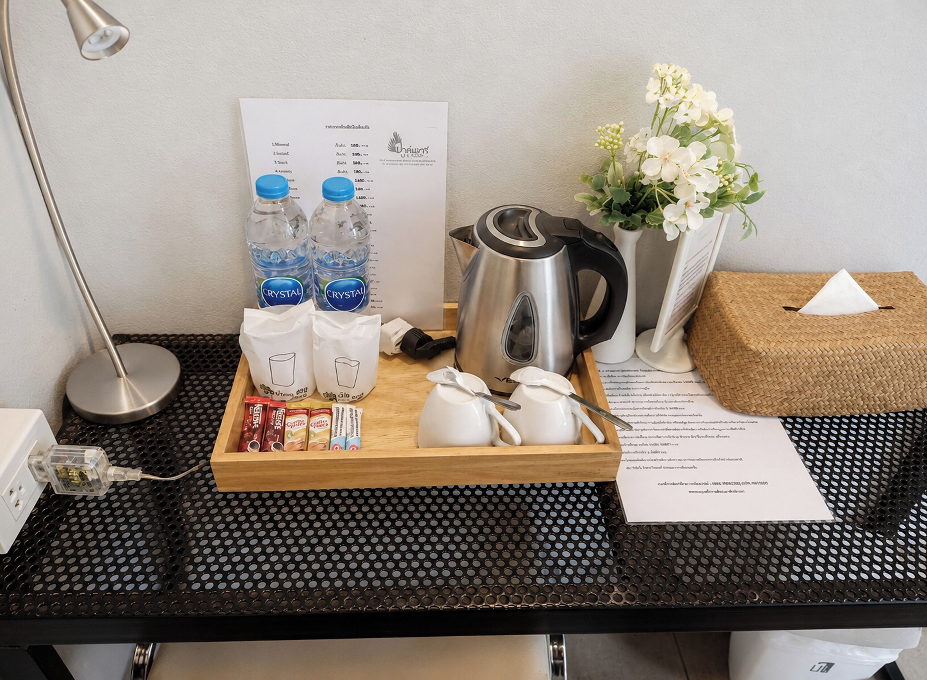 Sur le bureau de la chambre d’hôtel, on trouve un plateau contenant des bouteilles d’eau, des sachets de café, une bouilloire et une boîte de mouchoirs.