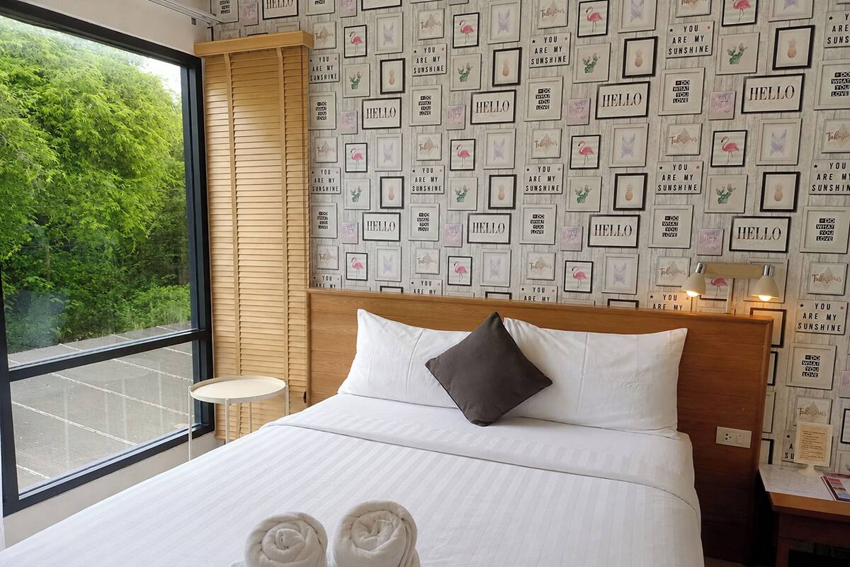 Een hotelkamer met een groot bed, een houten hoofdeinde, een kleine ronde tafel en uitzicht op groen door een groot raam.