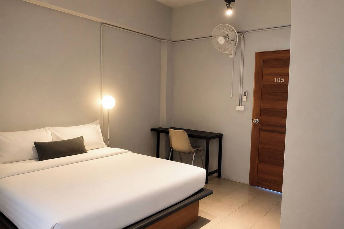 Een hotelkamer met een bed, een bureau, een stoel, een houten deur en een ventilator aan de muur.