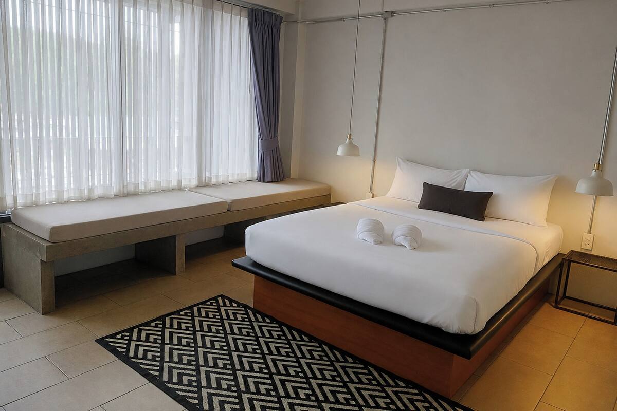 Een hotelkamer met een bed, een bankje, een kamerkleed met dessin en een raam met gordijnen.