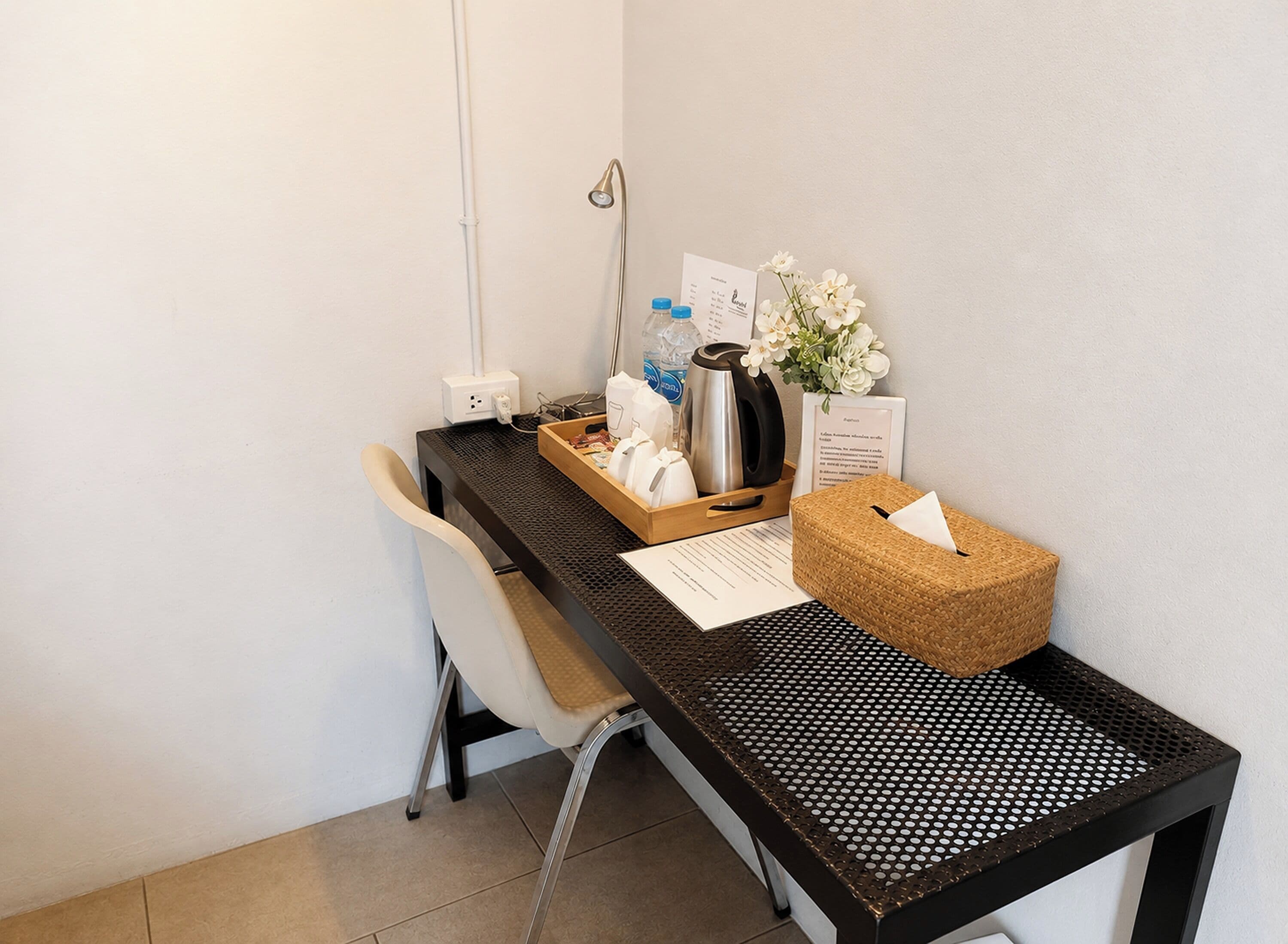 A hotel reception desk with a chair, a black metal desk, a white vase with flowers, a tray with toiletries, and a small wooden box.