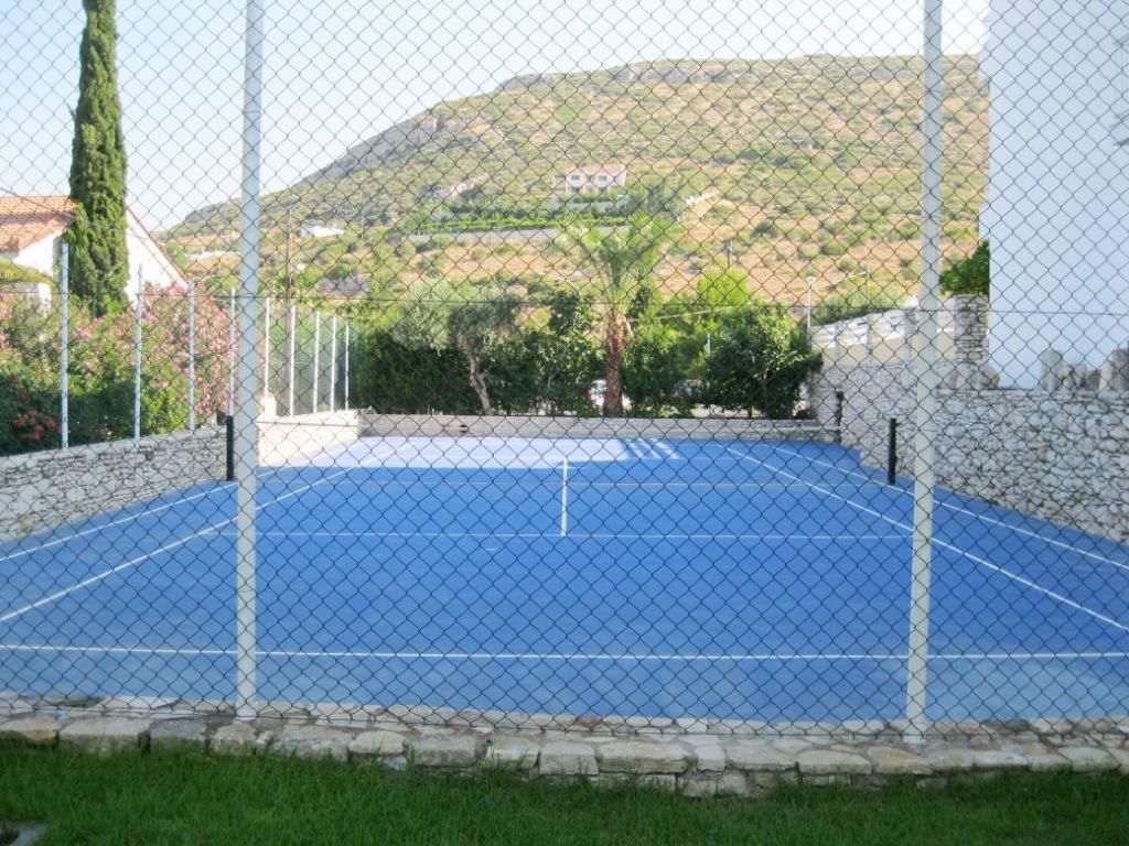 An outdoor tennis court with a chain-link fence, surrounded by stone walls and greenery.