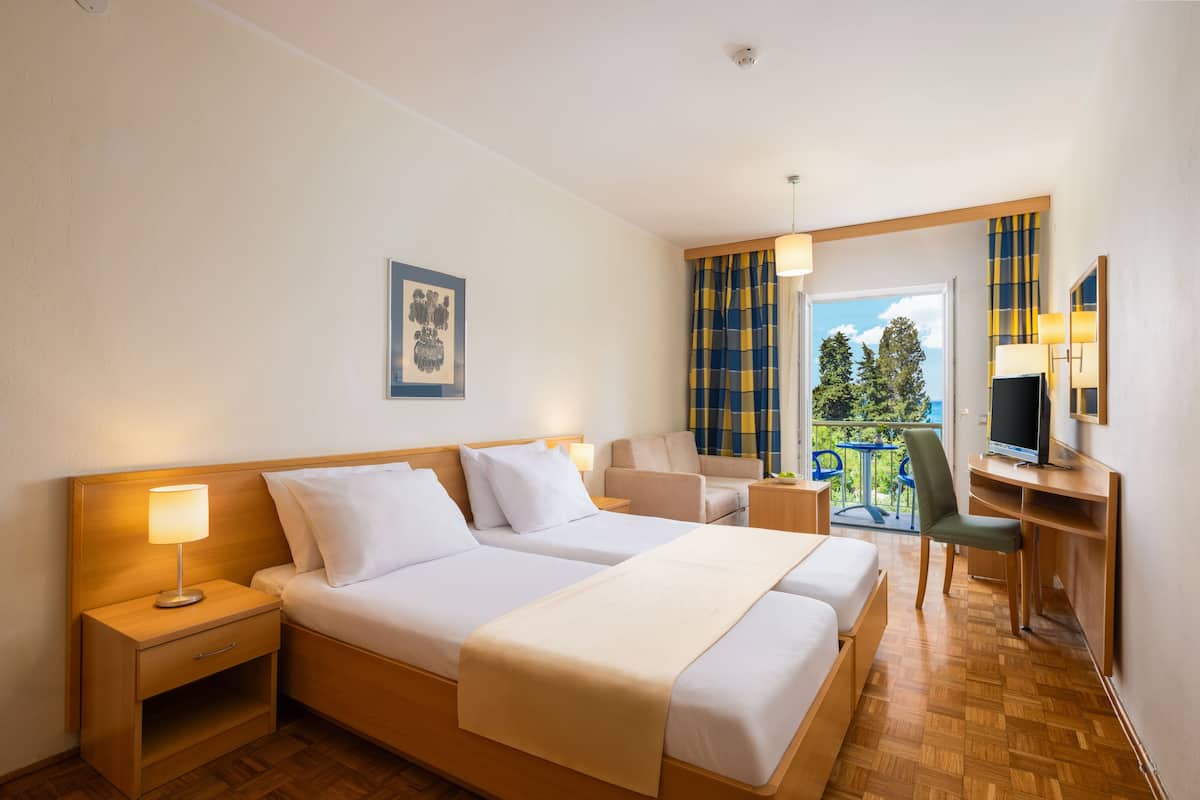 Habitación de hotel con dos camas, un escritorio con silla, televisión y balcón con vista a árboles.