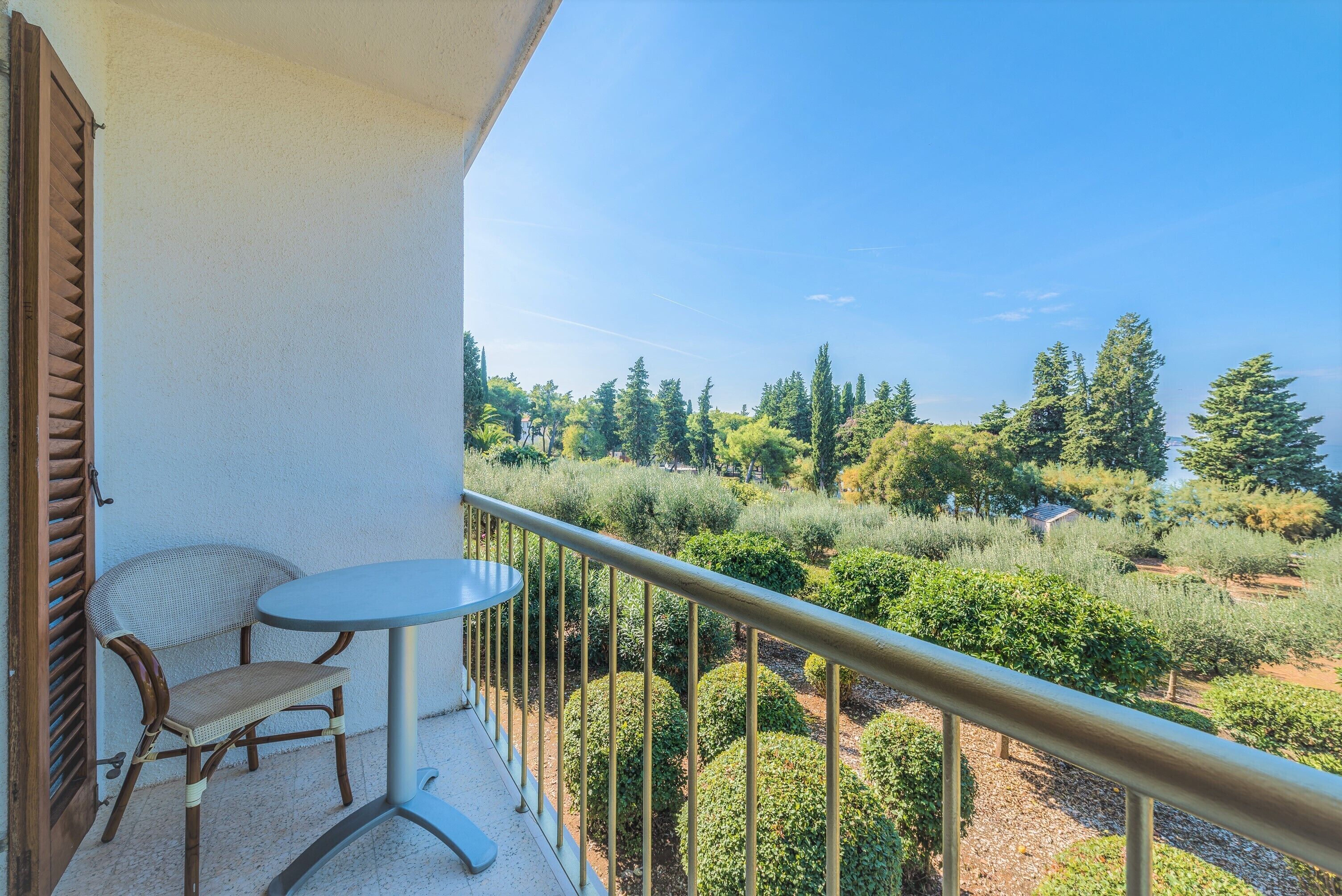 A balcony with a table and chairs, offering a view of a landscaped garden with various trees and shrubs.