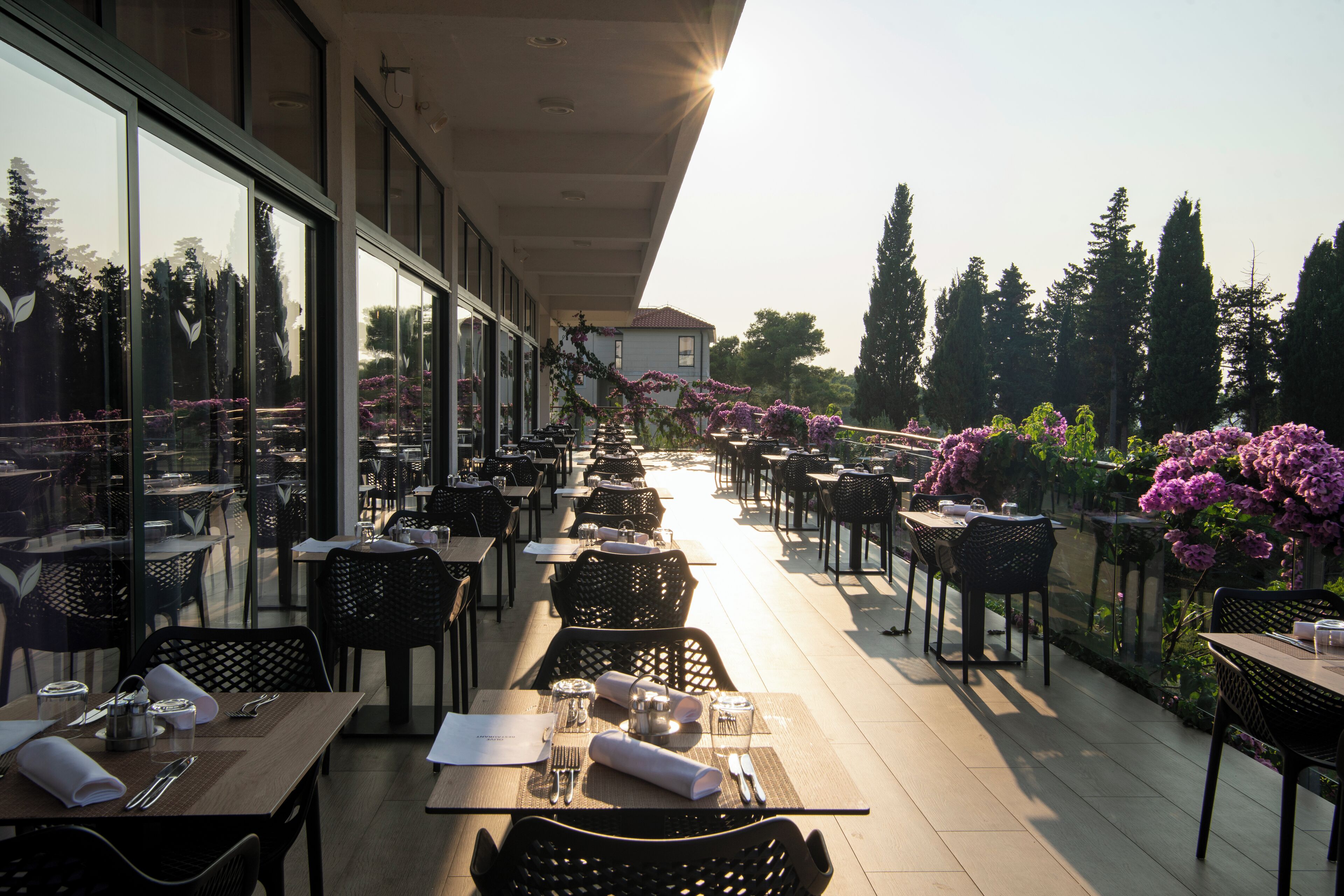 Un espace extérieur aménagé pour les repas, avec des tables et des chaises, de grandes fenêtres et une vue sur des arbres et un bâtiment en arrière-plan.