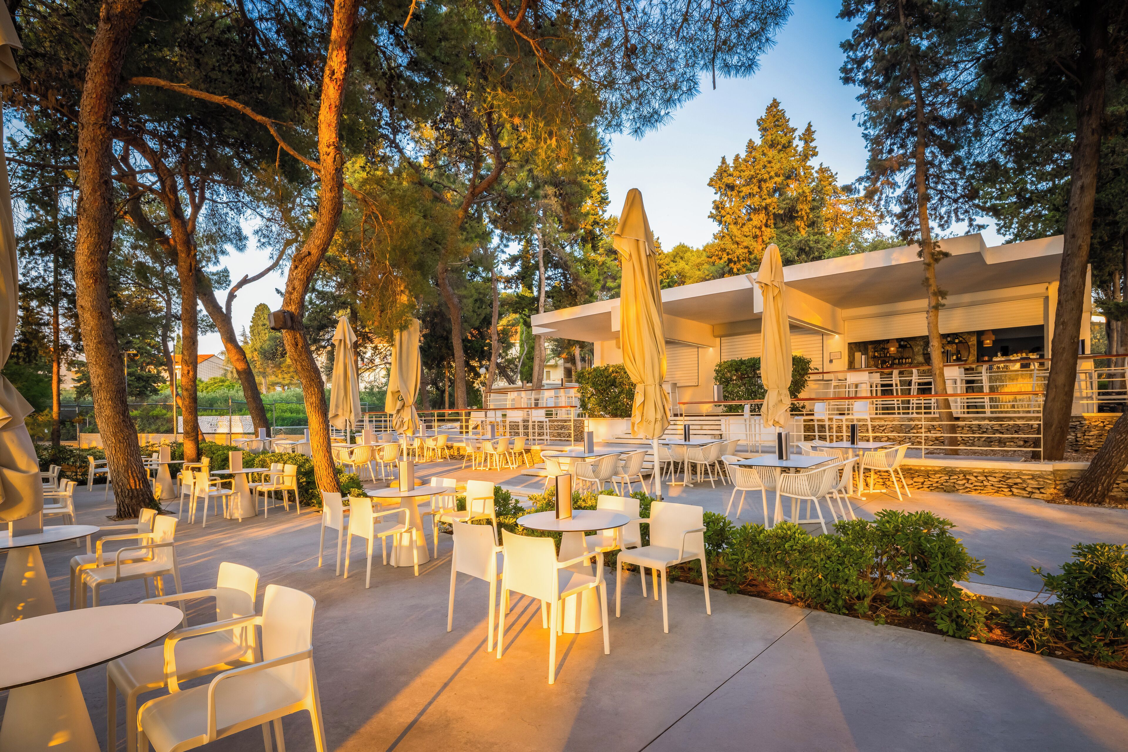 An outdoor patio area with round tables and white chairs, surrounded by trees and a modern building in the background.