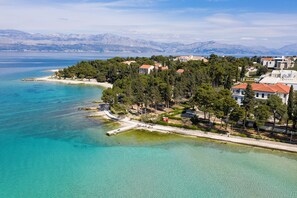 An aerial view of a coastal area with a beach, clear turquoise waters, and a cluster of buildings with red-tiled roofs. - Aminess Vival Casa Velaris  (Supetar)