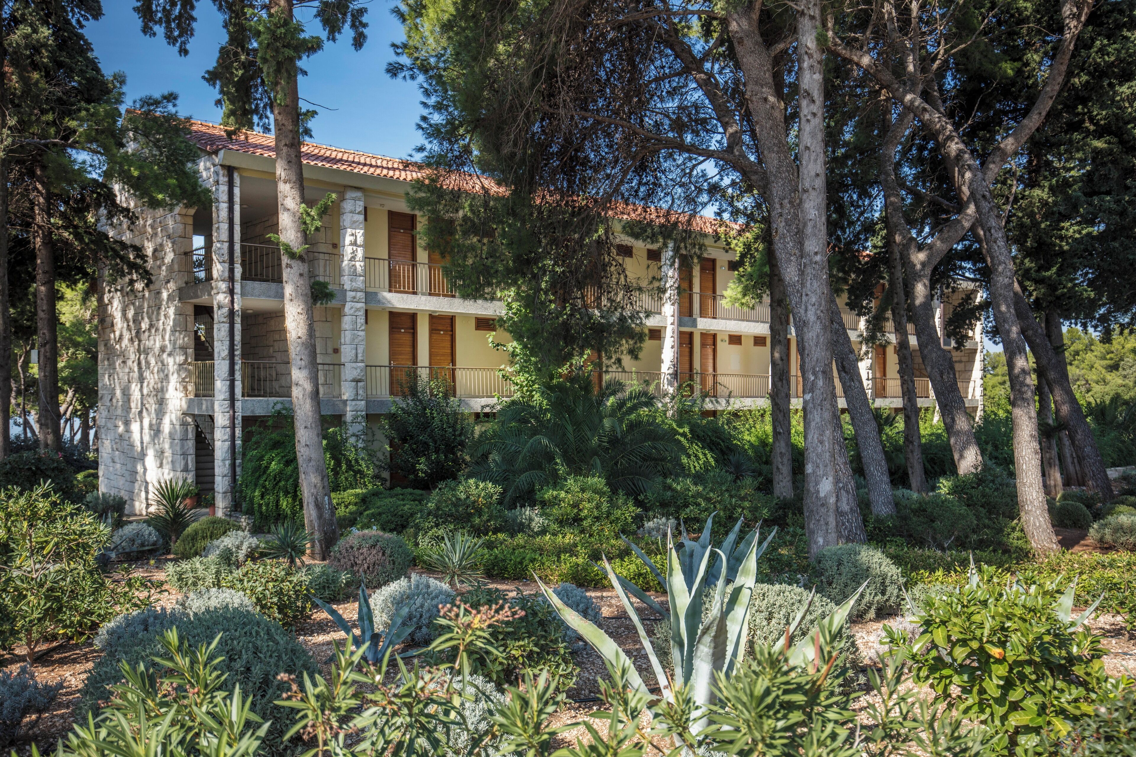 A multi-story building with balconies, surrounded by a variety of trees and shrubs.