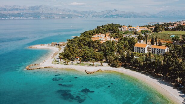 An aerial view of a coastal area with a beach, clear turquoise waters, and a mix of residential buildings with red roofs. - Aminess Vival Casa Velaris  (Supetar)