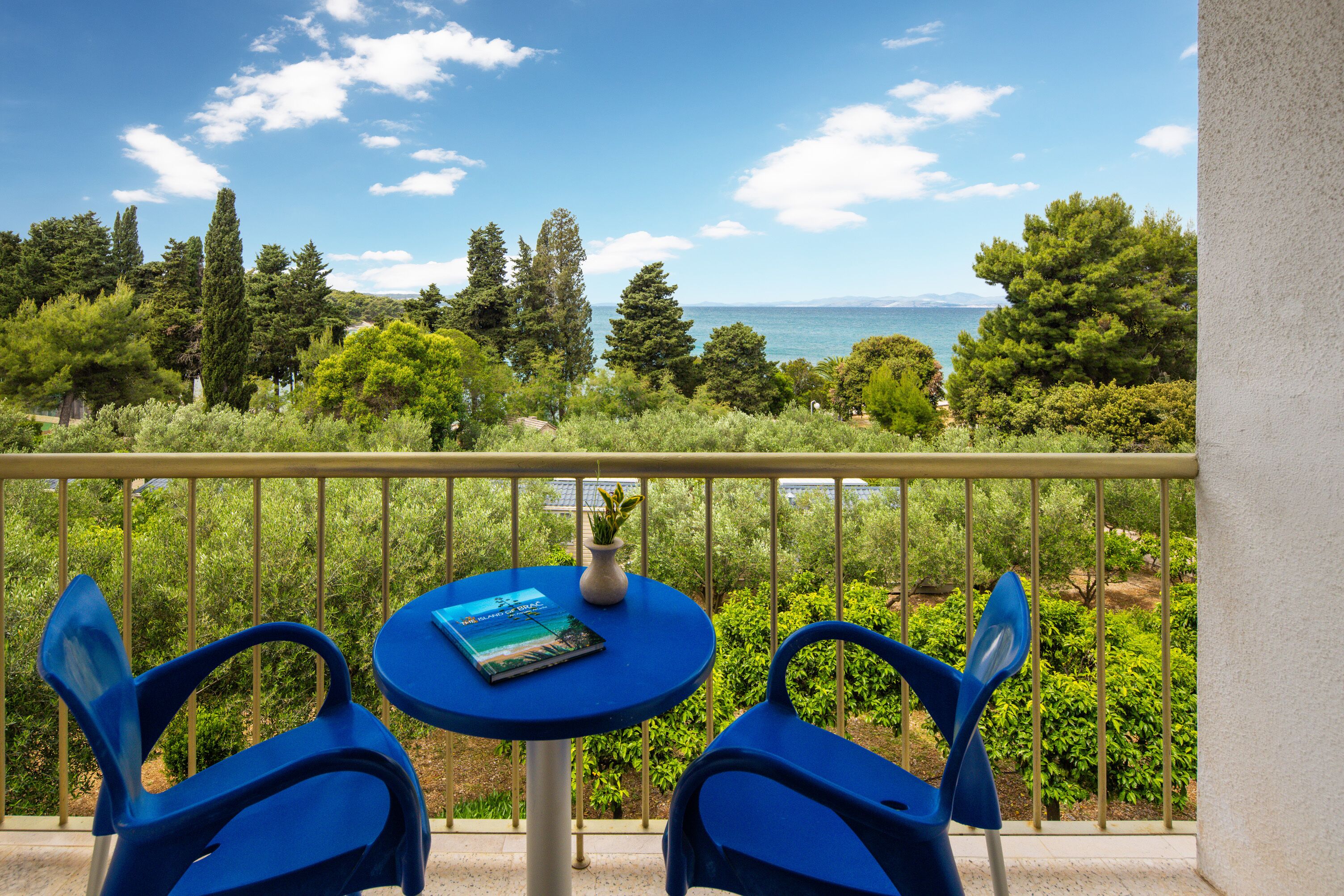 A balcony with two blue chairs and a small table, overlooking a scenic view of trees and the sea.