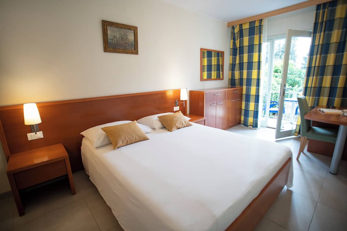 Habitación de hotel con una cama grande, cabecero de madera, dos mesitas de noche con lámparas, un armario de madera y vistas al exterior a través de un ventanal.