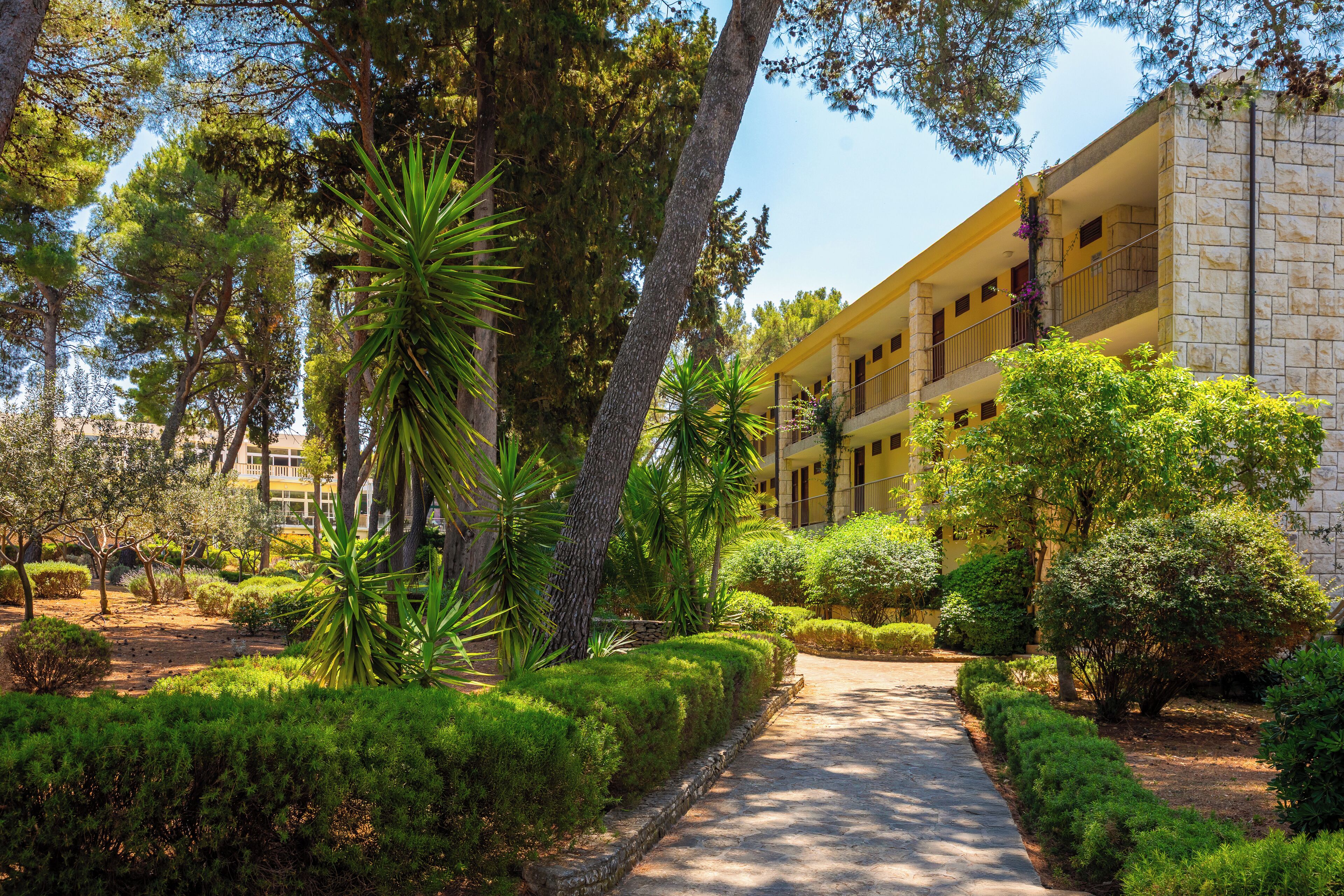 A building with a yellow facade, surrounded by well-maintained greenery and a paved pathway.