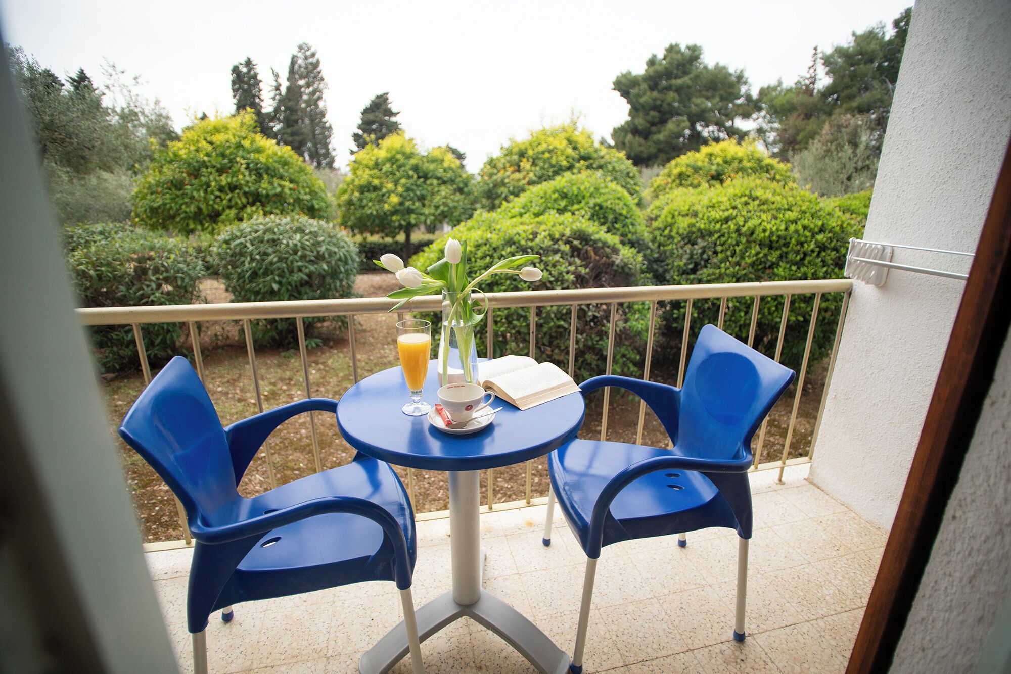 A small round table with two blue chairs on a balcony with a view of neatly trimmed bushes and trees.