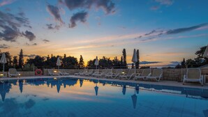 A swimming pool with lounge chairs and umbrellas, surrounded by trees and a sunset sky. - Aminess Vival Casa Velaris  (Supetar)