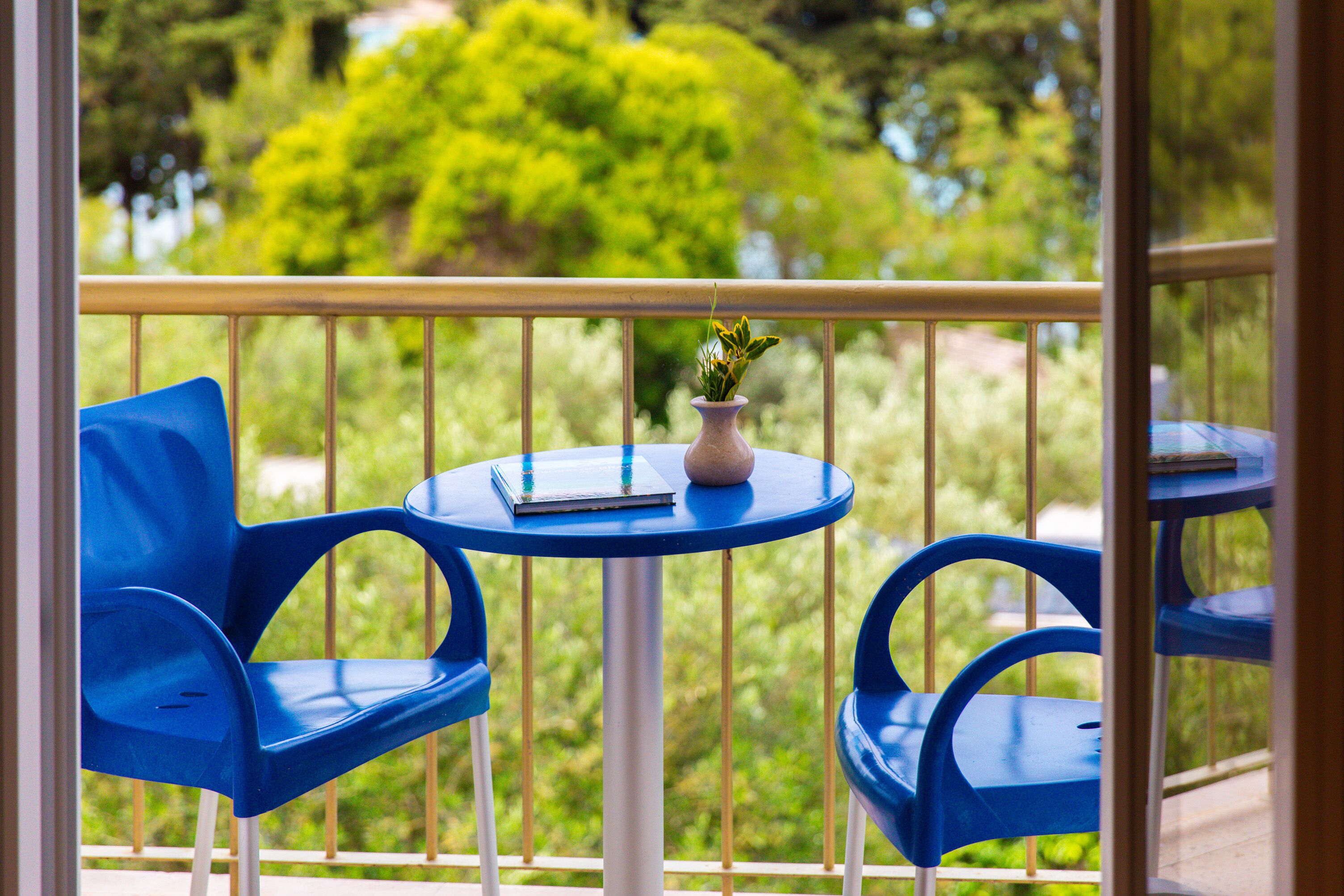 Ein Balkon mit zwei blauen Stühlen und einem kleinen Tisch, auf dem ein Buch und eine Vase stehen.