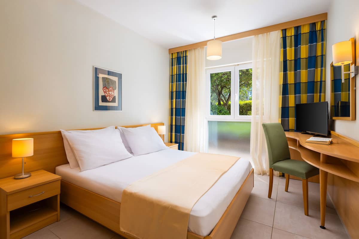 Una habitación de hotel con cama, escritorio, silla, televisor y ventana con cortinas.