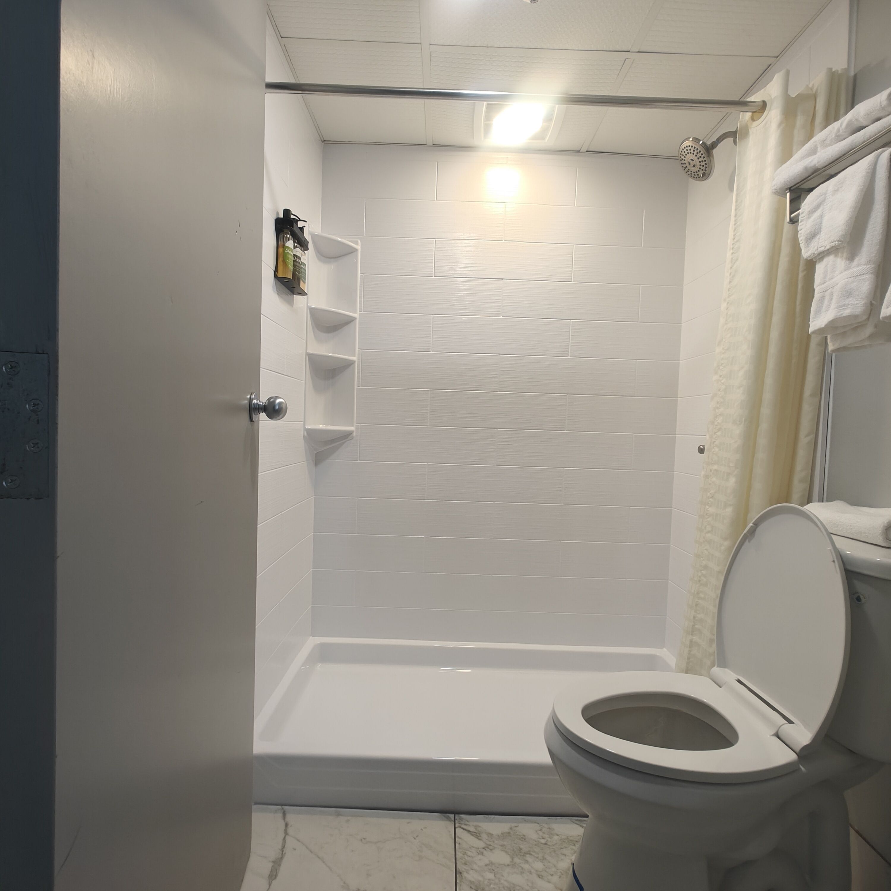 A bathroom with a white tiled shower, a white toilet, and a shelf with toiletries.