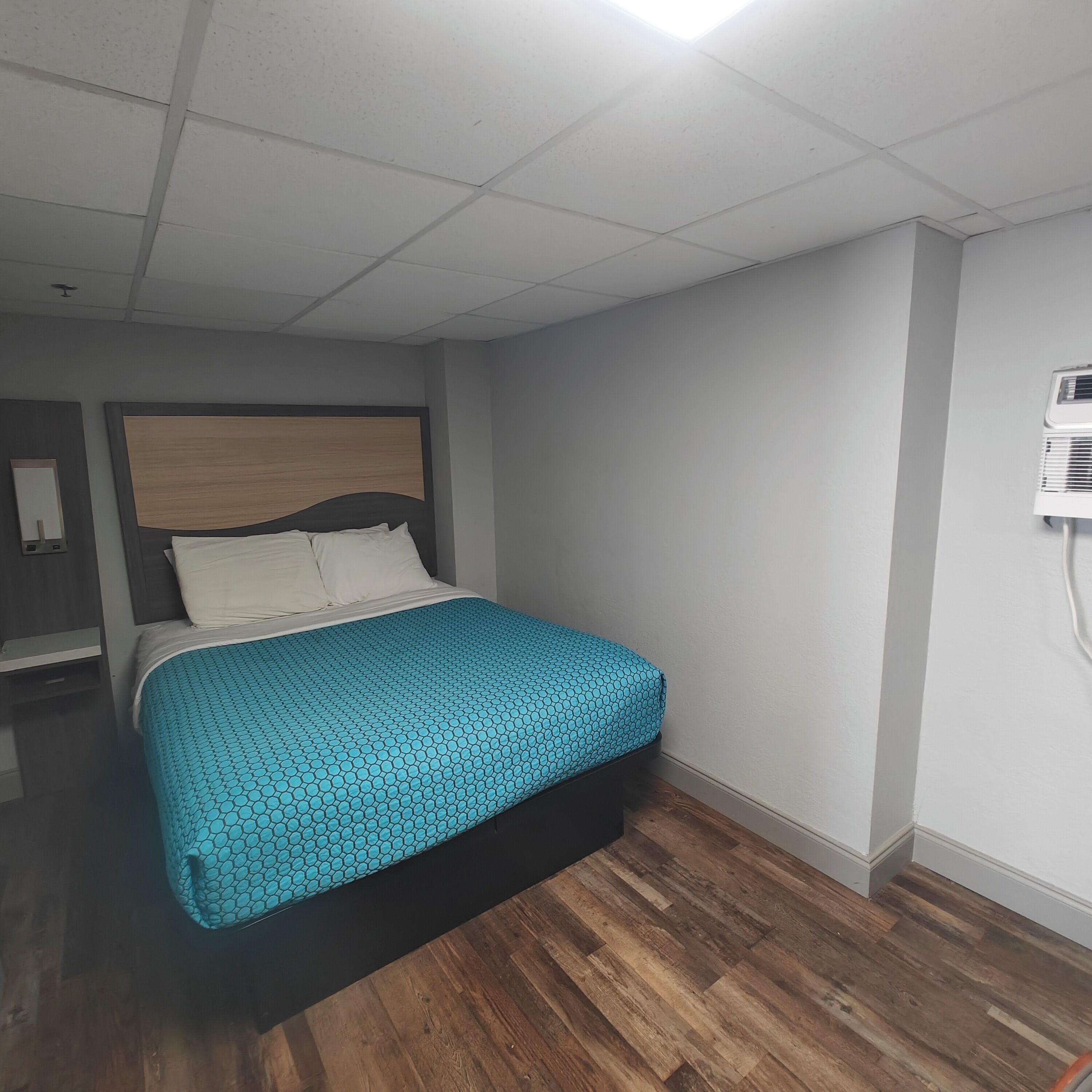 A hotel room with a bed, wooden flooring, a headboard, and a wall-mounted air conditioning unit.