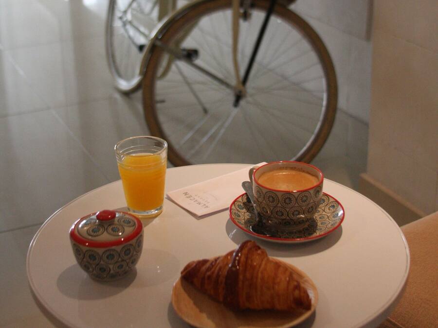 Een ontbijttafel met een croissant, een kop koffie, een glas sinaasappelsap en een klein bakje, met een fiets op de achtergrond.