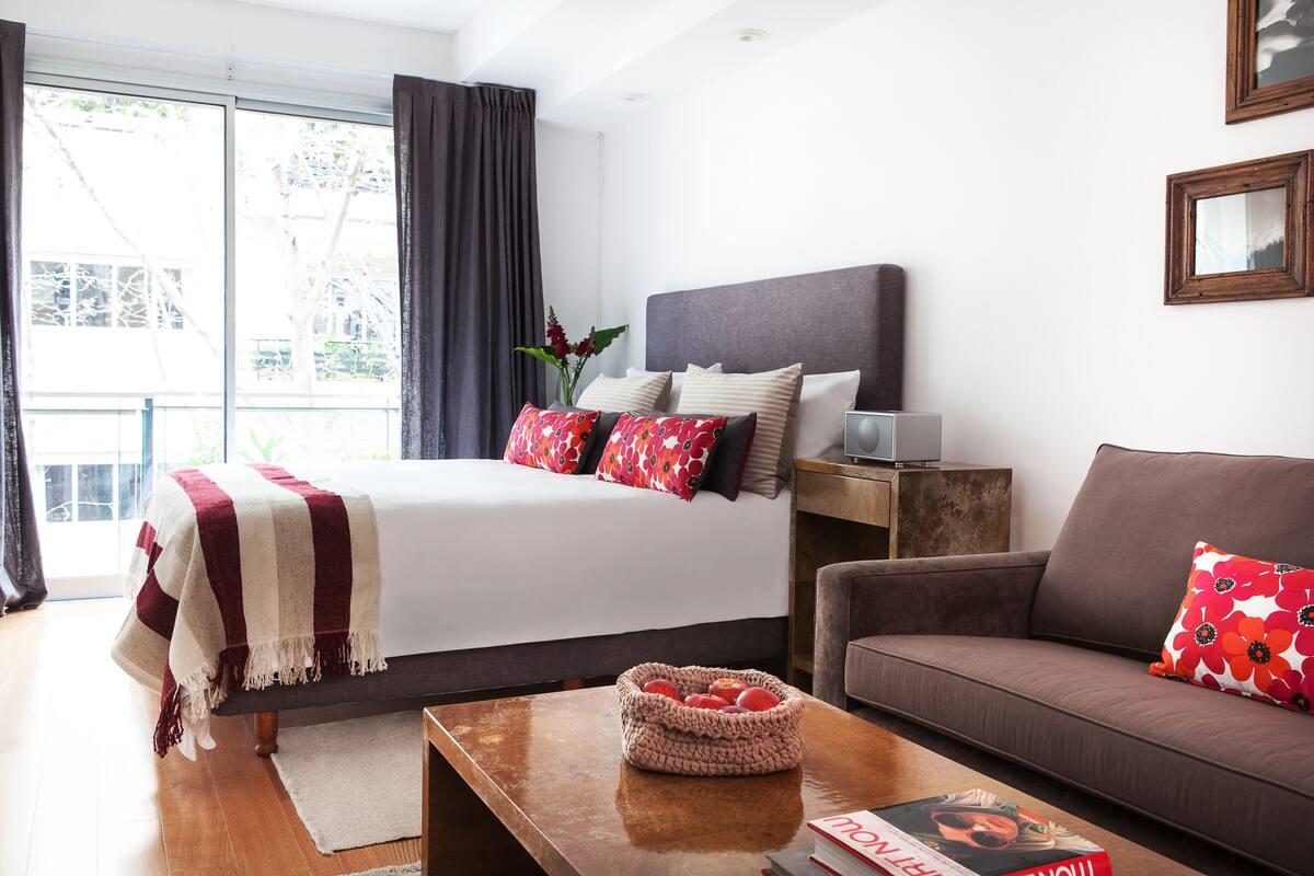A modern hotel room with a large bed, a sofa, a coffee table, and a window with curtains.