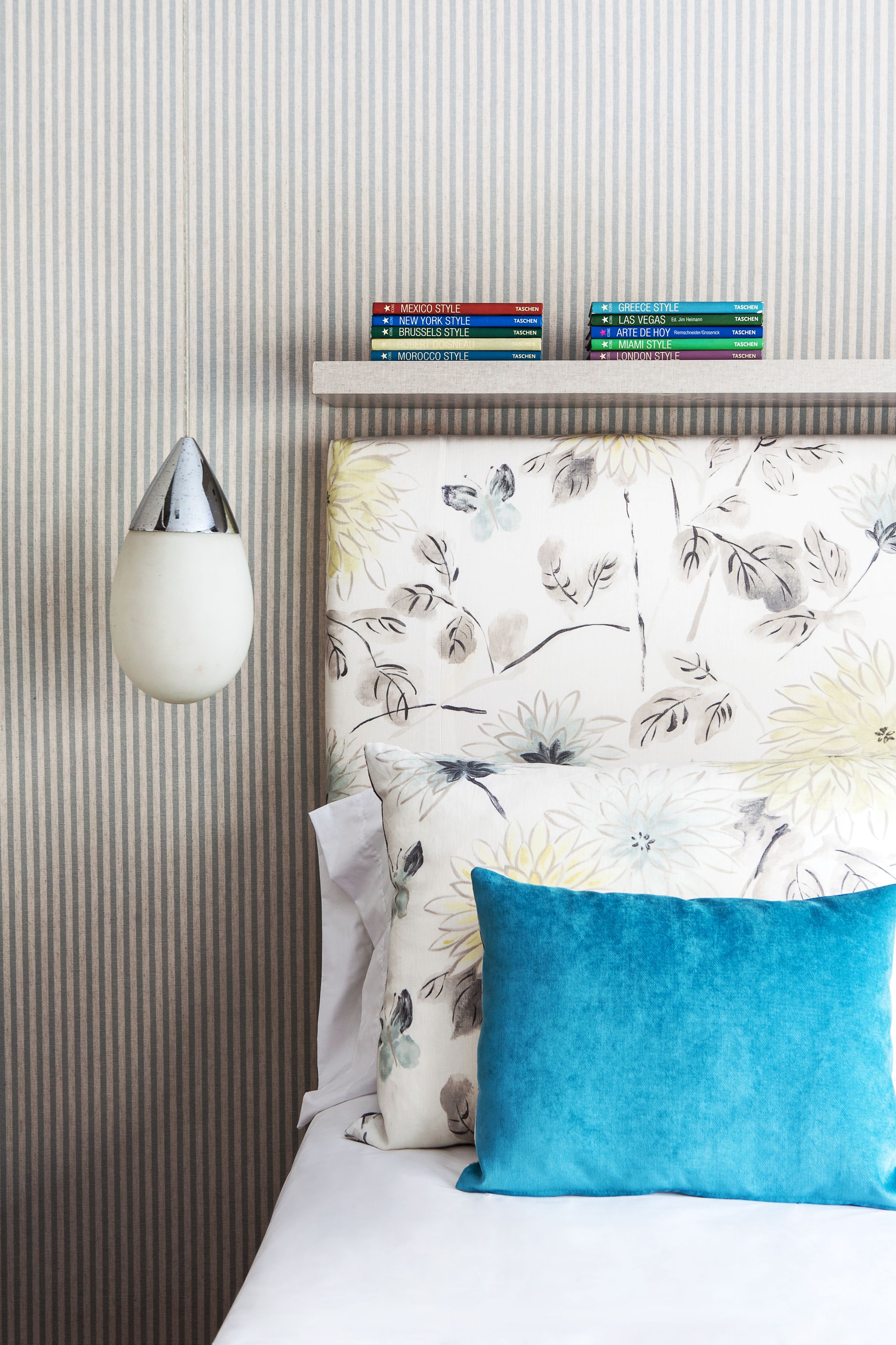 Un cuarto de hotel ordenado con papel tapiz a rayas, cabecera con estampado floral, lámpara de noche blanca y una selección de libros en un estante.
