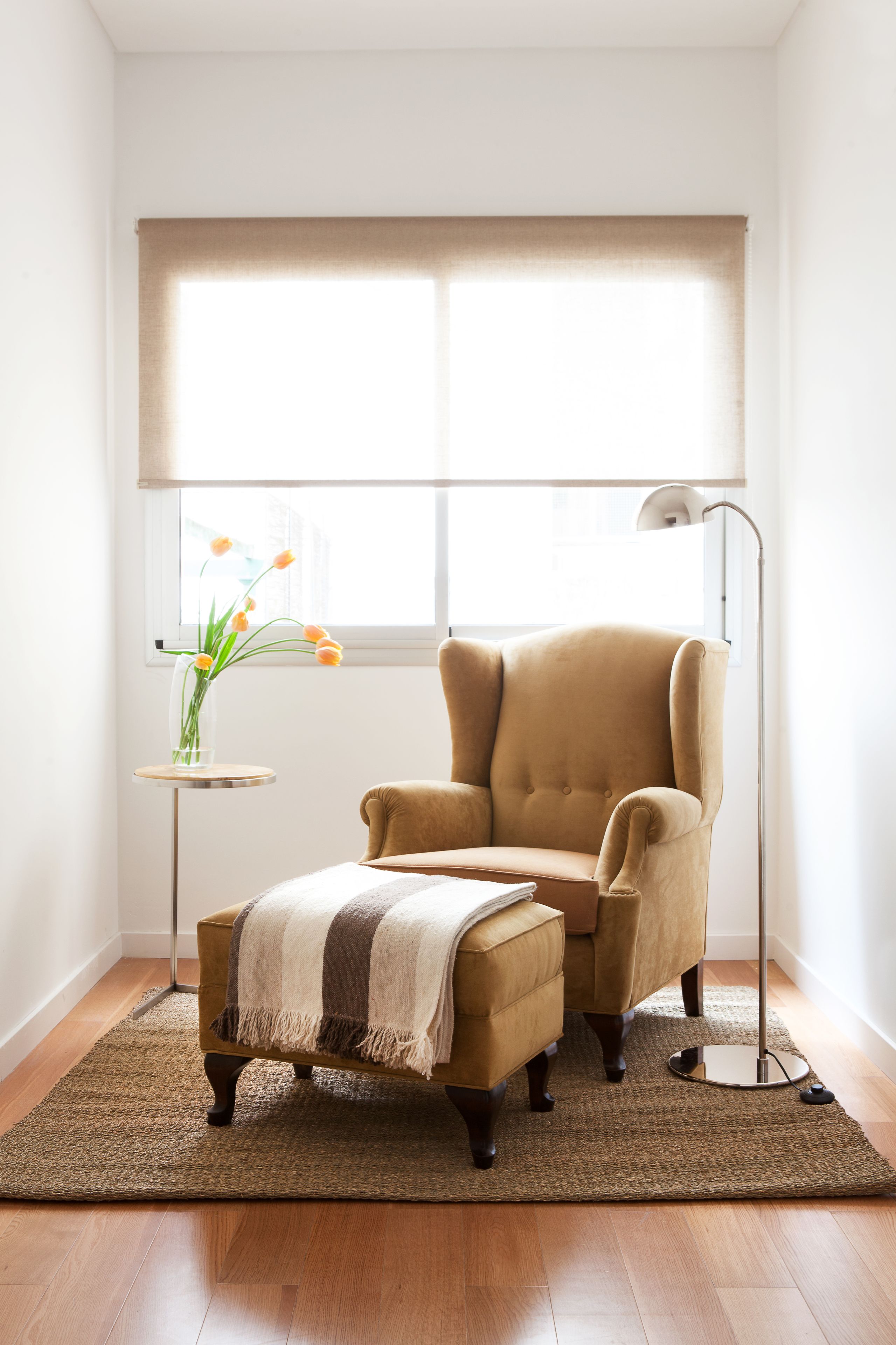A room with a large window, a beige armchair, a small side table with a vase of flowers, and a floor lamp.