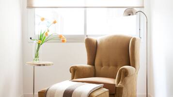 A room with a large window, a beige armchair, a small side table with a vase of flowers, and a floor lamp.