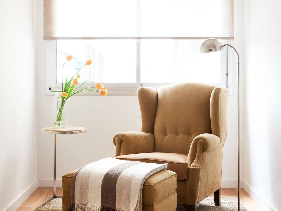 A room with a large window, a beige armchair, a small side table with a vase of flowers, and a floor lamp.