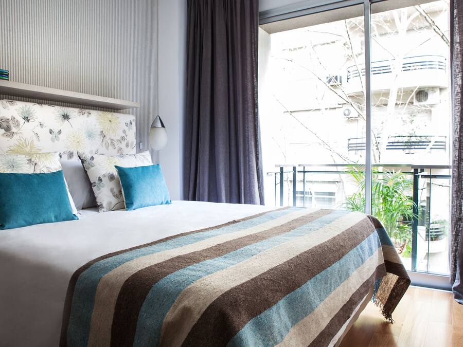 A hotel room with a large bed, a view of the city through the window, and a balcony with plants.