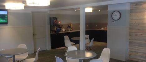 A modern dining area with round tables and white chairs, a kitchen in the background, and a clock on the wall.