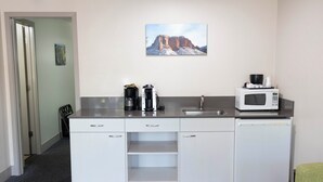 Une cuisine compacte équipée d’un four à micro-ondes, d’une machine à café et d’un lave-vaisselle. Un tableau orne le mur, et une porte donne accès à une autre pièce.