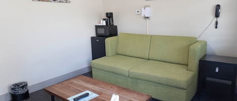 A room with a green sofa, a wooden coffee table with a remote control and tissues, a black side table, a black speaker, a black microwave, and a trash bin.